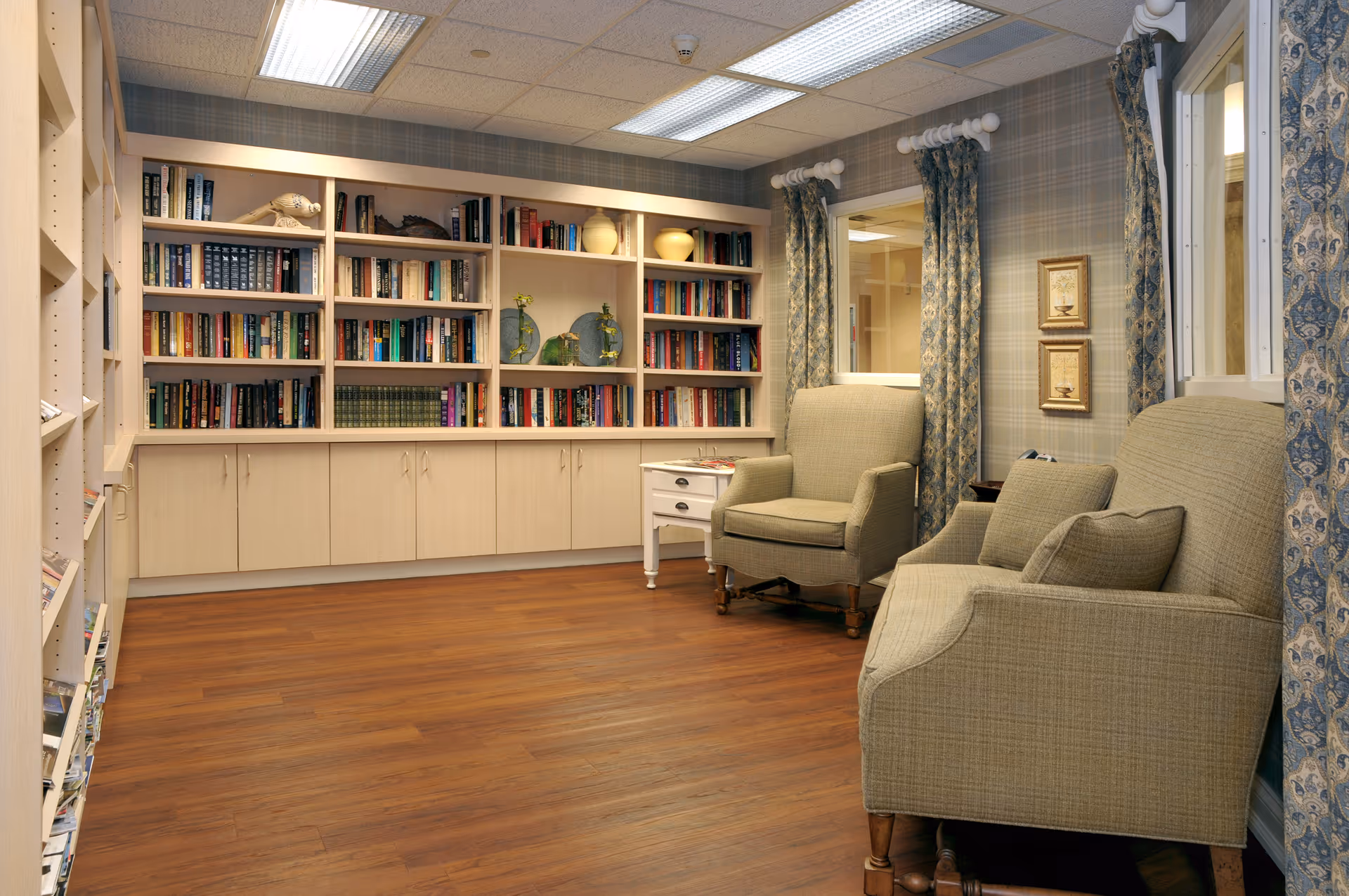 A cozy reading room with wooden floors, built-in bookshelves filled with books and decorative items, two upholstered armchairs, a small white side table, and windows with patterned curtains.