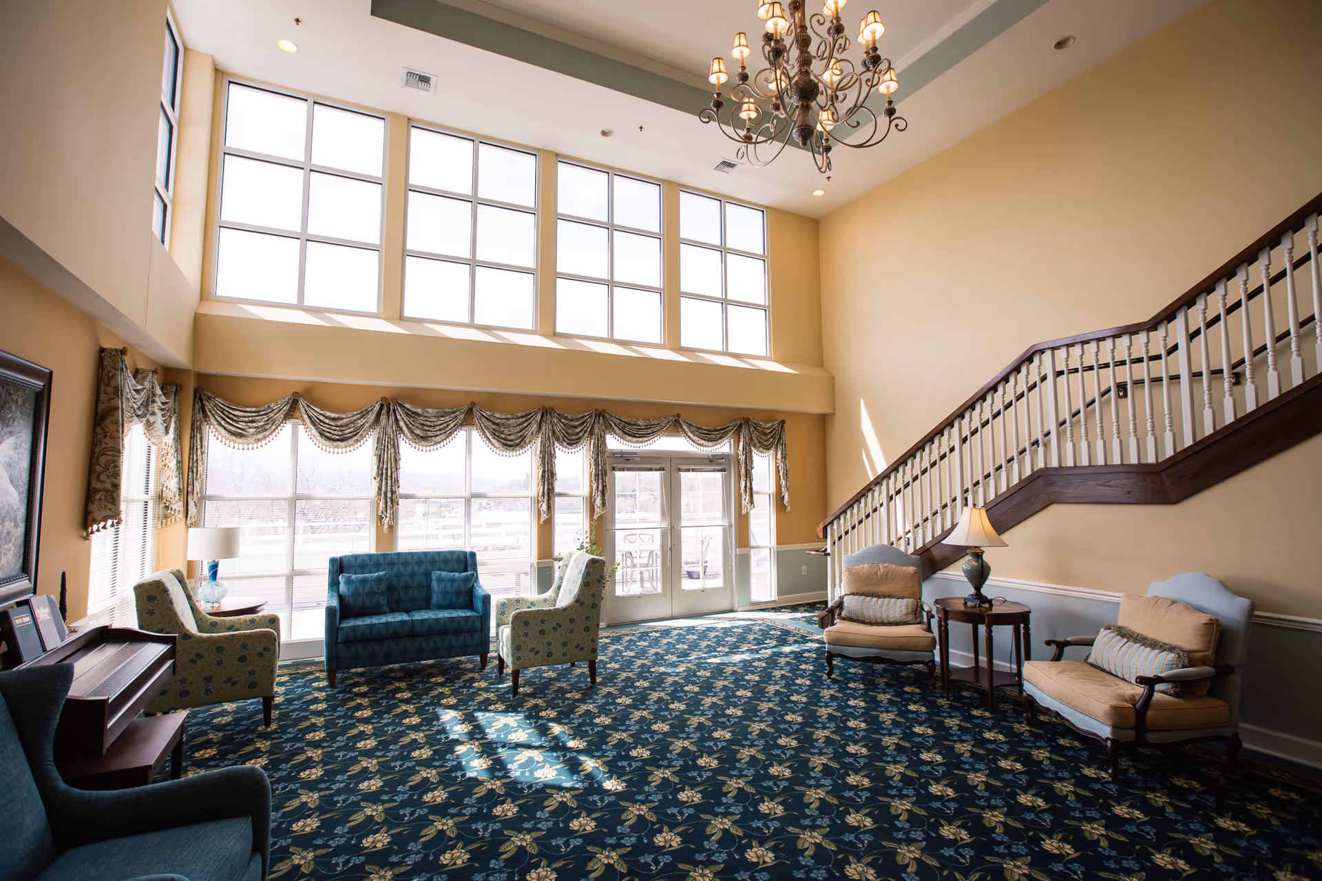 A spacious, well-lit living room area with large windows letting in natural light. The room features a patterned blue carpet, several upholstered chairs and sofas, a small side table with a lamp, a piano, and a staircase with wooden railing leading to an upper floor.