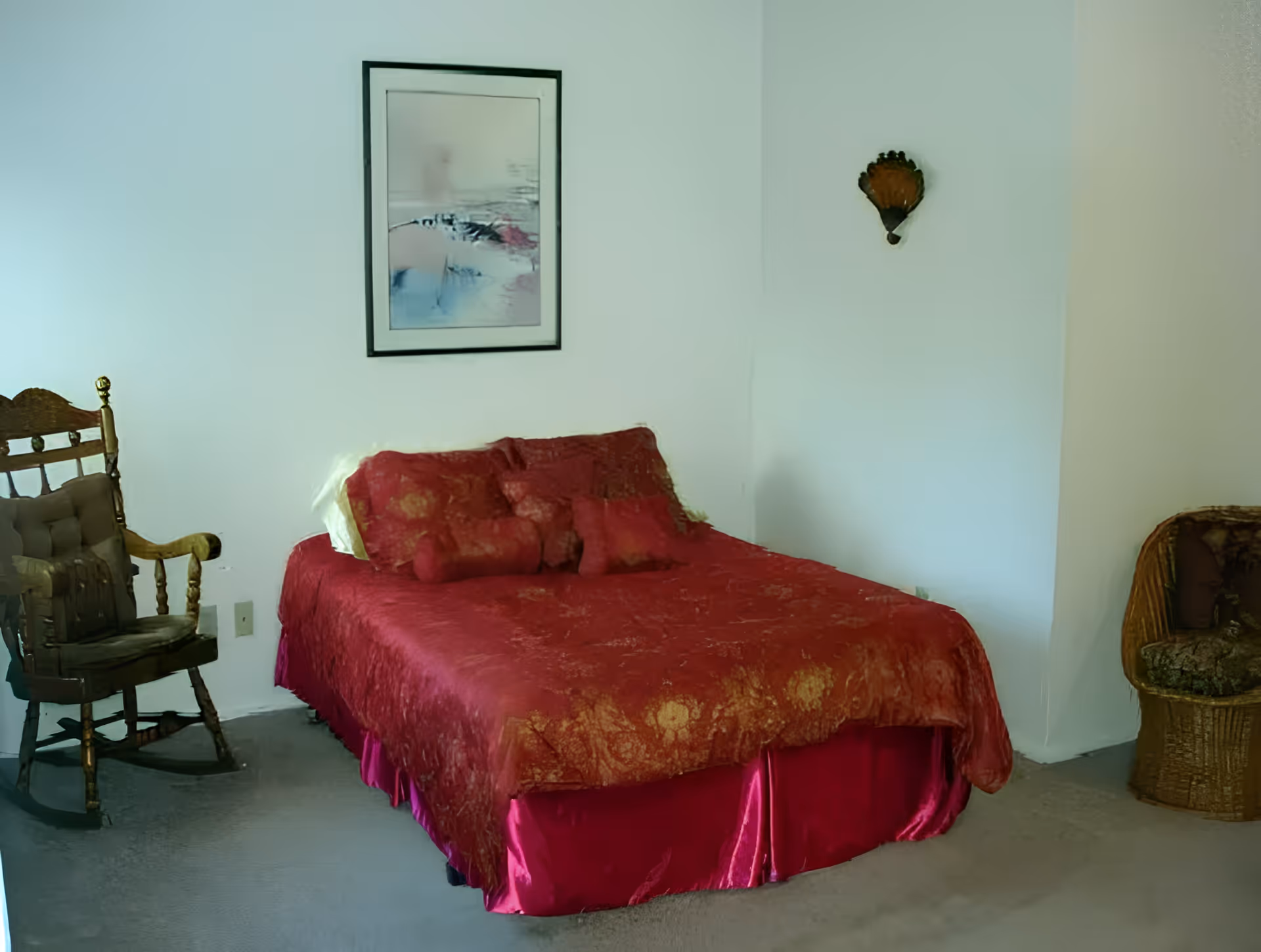 A bedroom with a bed covered in a red and gold patterned bedspread and matching pillows. To the left of the bed is a wooden rocking chair with a cushion, and to the right is a wicker chair with a cushion. A framed abstract painting hangs on the wall above the bed, and a small decorative wall hanging is on the adjacent wall.