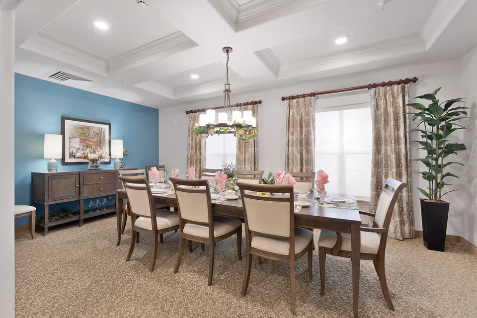 A well-lit dining room with a long wooden table set for eight people. The table is decorated with pink napkins, plates, and glassware. There are eight cushioned chairs around the table. The room has beige patterned carpet, two large windows with patterned curtains, a blue accent wall with a framed painting, two table lamps on a wooden sideboard, and a tall potted plant in the corner. A chandelier with multiple lights hangs above the table.