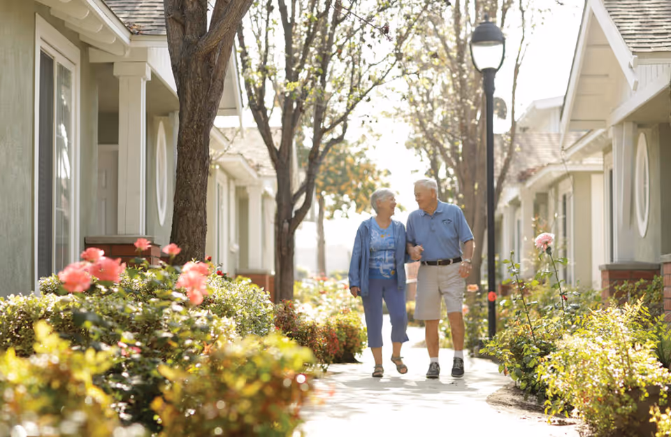 An elderly couple walking hand in hand along a garden pathway lined with blooming flowers and trees, with residential buildings on either side.