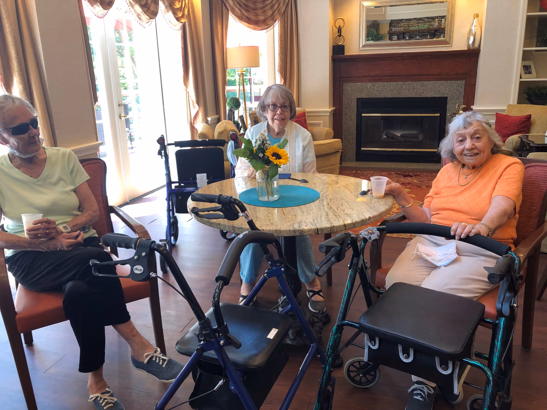 Three elderly women sitting around a round table in a cozy living room area with a fireplace in the background. Two women are seated in chairs, and one woman is seated in a wheelchair. There are walkers positioned near the table, and a vase with flowers is placed on the table. The room has large windows with drapes, letting in natural light.