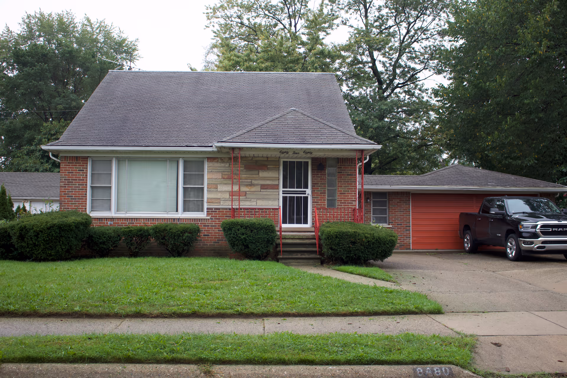 Front exterior view of a single-story brick house with a gray shingled roof, a small porch with red railings, a large window with closed blinds, green bushes in front, a driveway leading to a garage with a red door, and a black pickup truck parked in the driveway. Trees are visible in the background.