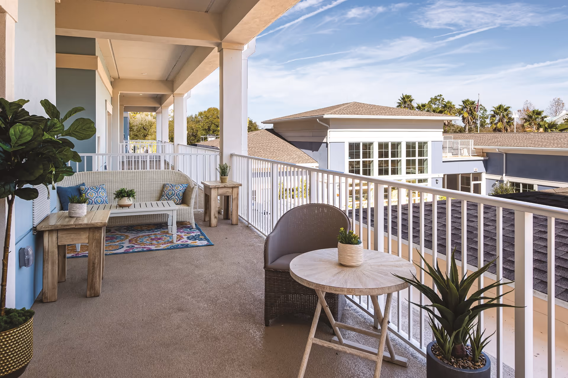 A spacious outdoor balcony area at Sancerre at Palm Coast with a white wicker loveseat adorned with blue patterned cushions, a small wooden coffee table, two wooden side tables with potted plants, a round wooden table with a wicker chair, and several potted plants. The balcony overlooks nearby buildings with large windows and a clear blue sky above.