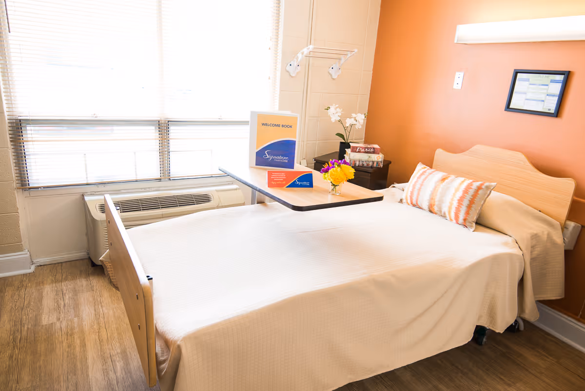 A neatly made single bed with a light wooden headboard and a beige bedspread in a room with an orange accent wall. There is a decorative pillow with orange and white stripes on the bed. A small overbed table holds a welcome book, a small bouquet of yellow flowers, and some informational cards. Next to the bed is a nightstand with a white orchid plant and a box of tissues. A large window with blinds and an air conditioning unit is visible on the left side of the room. The floor is wood-style laminate.