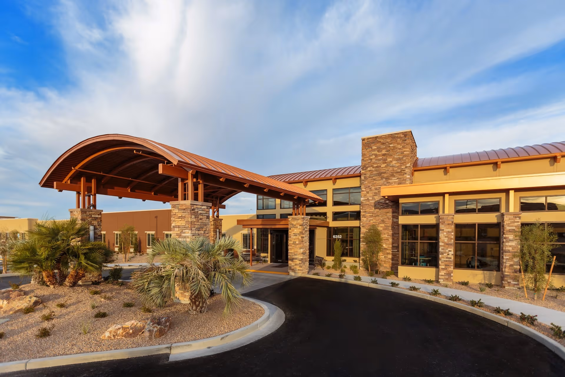 Exterior view of Canyon Vista Post-Acute facility featuring a covered entrance with stone pillars, desert landscaping with palm trees and rocks, and a clear blue sky with some clouds.