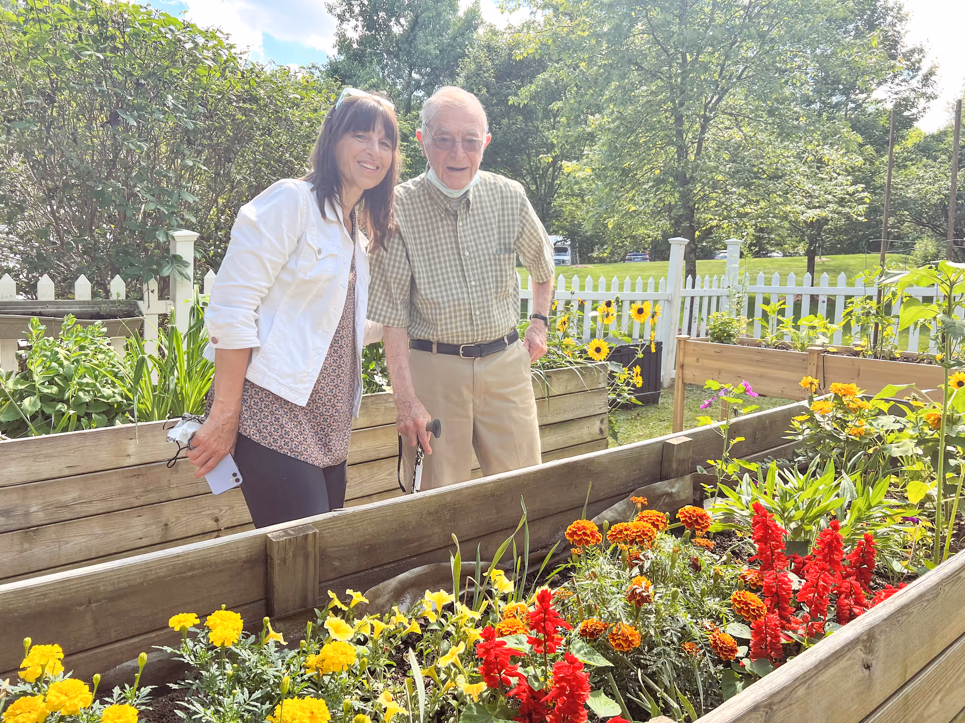 An elderly man and a middle-aged woman standing together outdoors in a garden area with raised wooden flower beds filled with colorful flowers including yellow, orange, and red blooms. They are smiling and enjoying the sunny day with green trees and a white picket fence in the background.