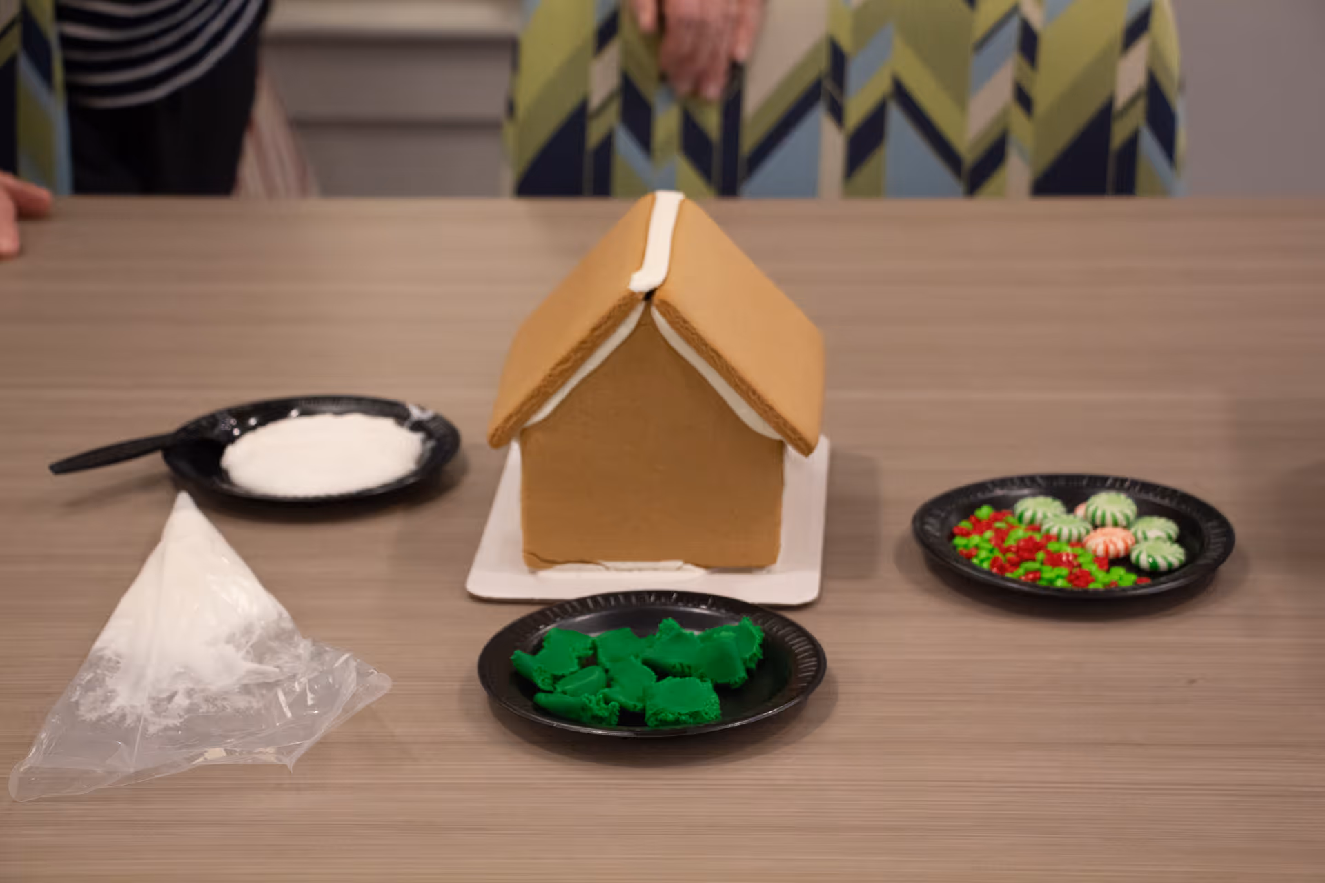 A gingerbread house kit on a table with a plain gingerbread house, a piping bag filled with white icing, and three black plates containing white icing, green icing shaped like trees, and red and white peppermint candies. Two people are partially visible behind the table.