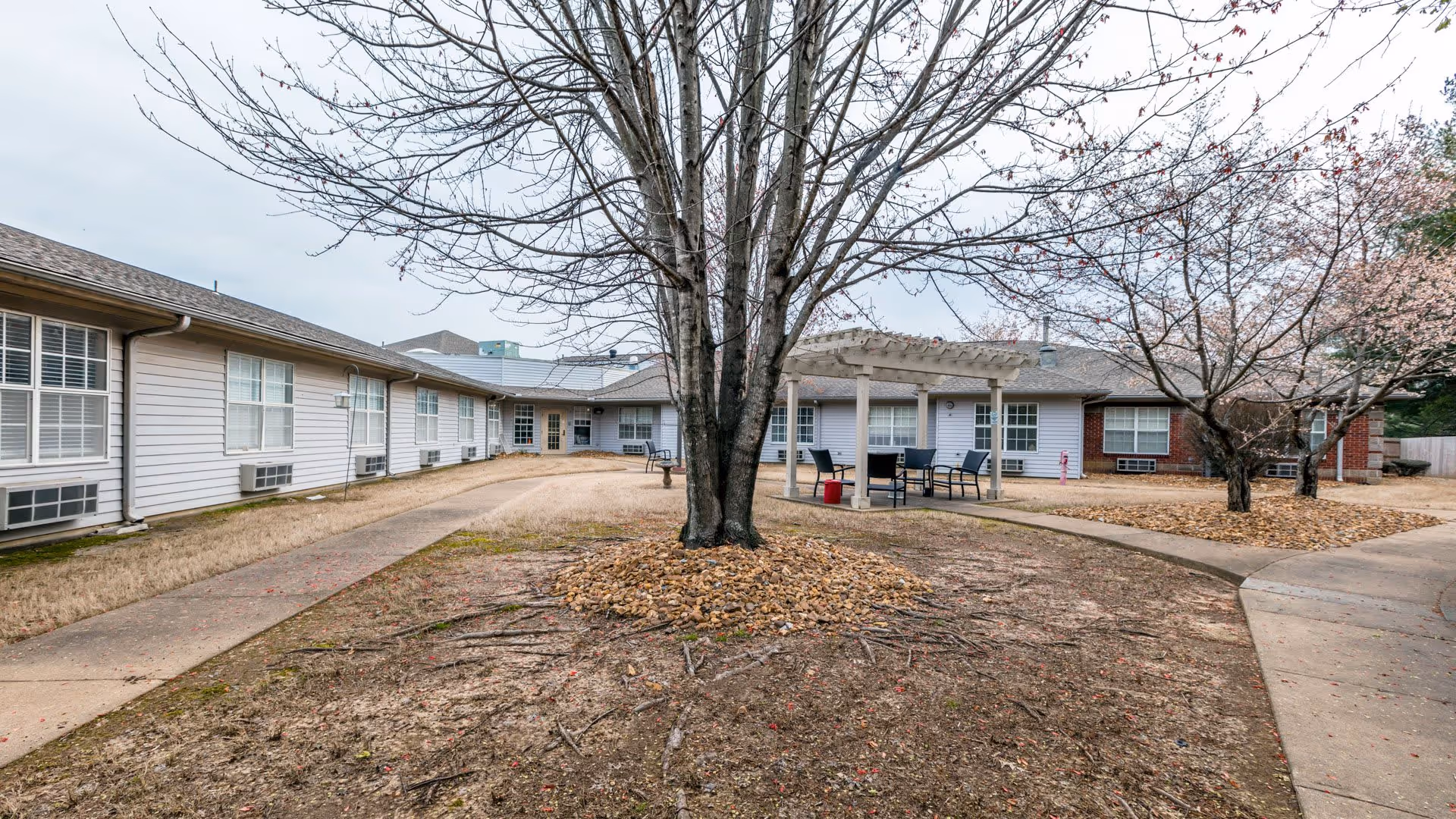 Outdoor courtyard area of a senior living facility with leafless trees, a pergola with chairs and a table underneath, surrounded by a single-story building with white siding and multiple windows.