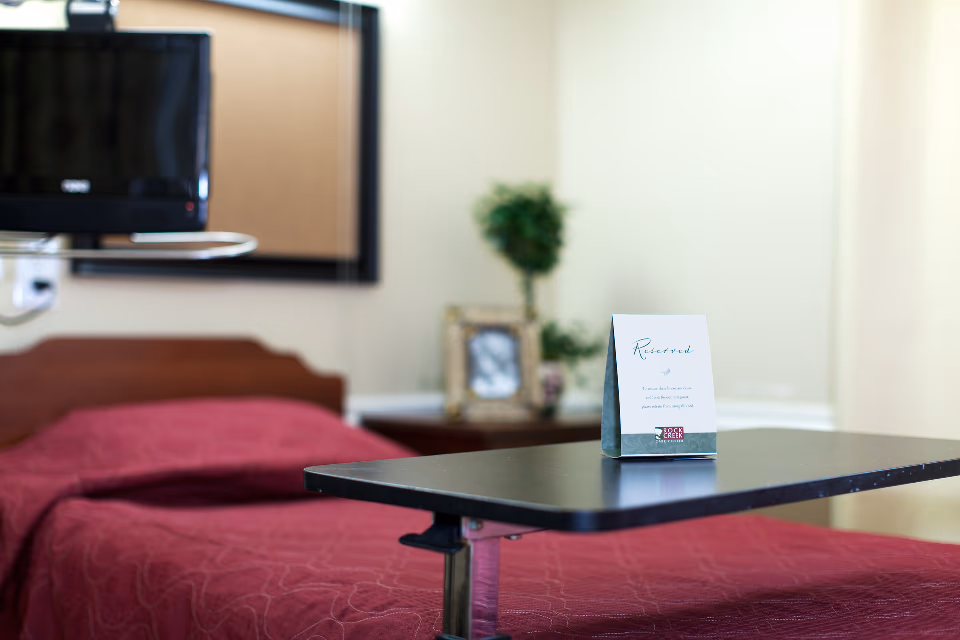 A single bed with a red bedspread in a room, featuring a black overbed table with a reserved sign on it. In the background, there is a small TV mounted on the wall, a framed photo, and a green plant on a side table.