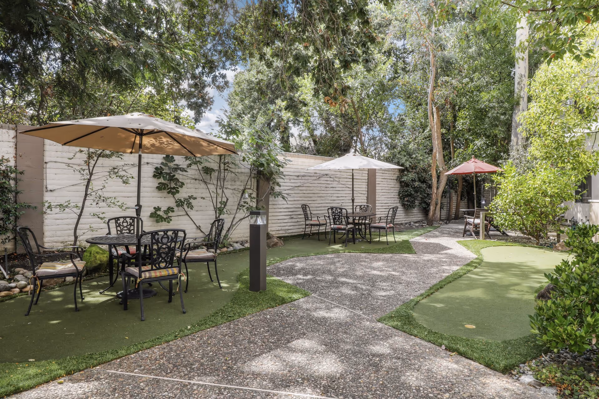 Outdoor patio area with several metal tables and chairs, each shaded by large umbrellas. The space is surrounded by trees and greenery, with a white wooden fence in the background and a paved walkway running through the area.