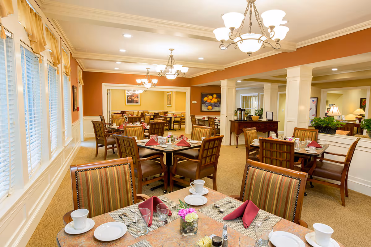 Bright, formal dining room with multiple set tables, upholstered chairs, and chandeliers.
