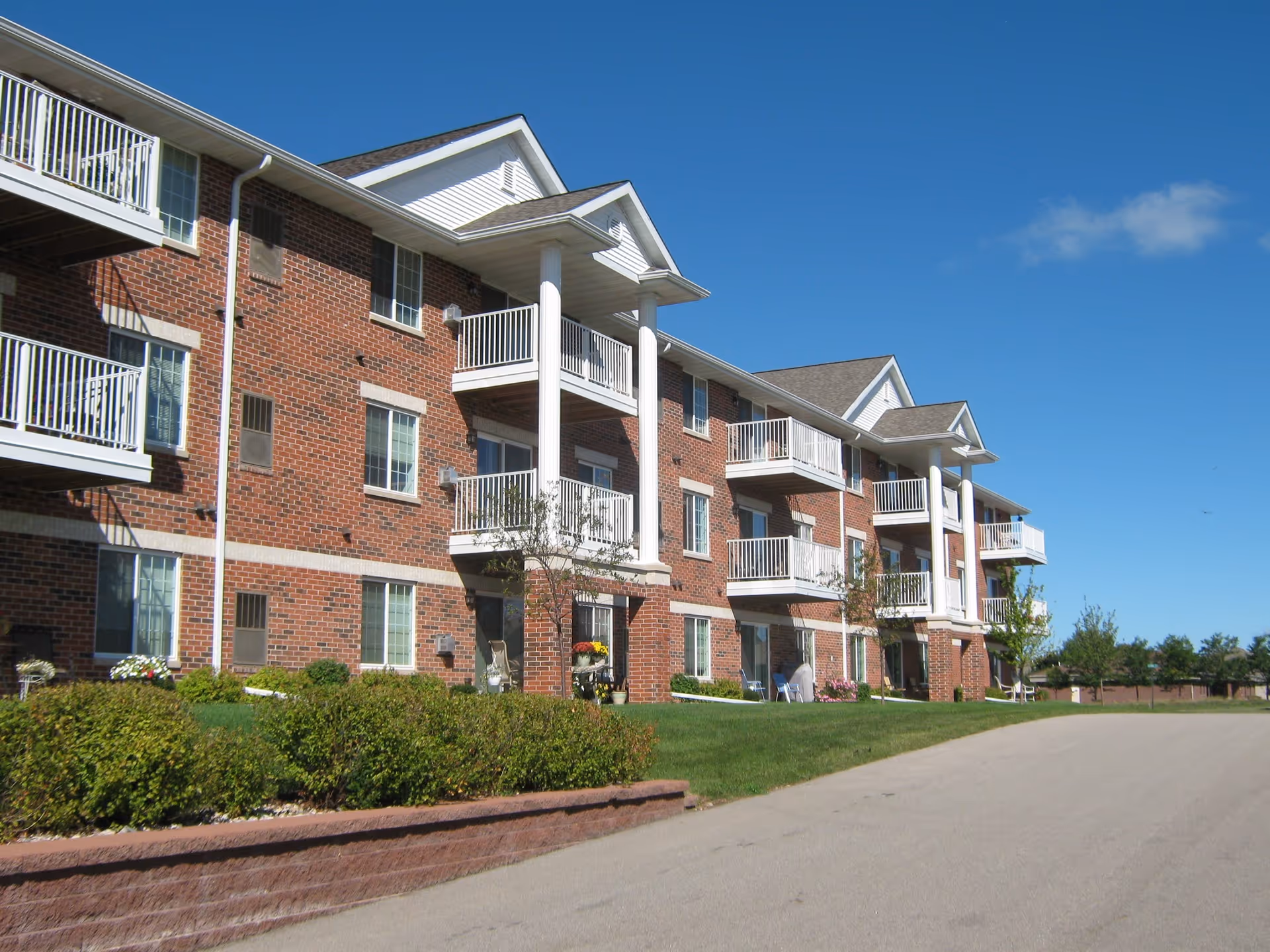 Exterior view of a three-story brick apartment building with white balconies and columns under a clear blue sky. The building is surrounded by green bushes and a well-maintained lawn next to a paved driveway.