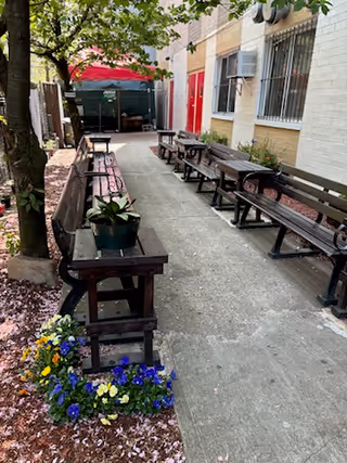 Outdoor seating area with multiple wooden benches and tables arranged along a concrete pathway next to a building. There are trees providing shade, flower beds with colorful flowers, and a red canopy visible in the background.
