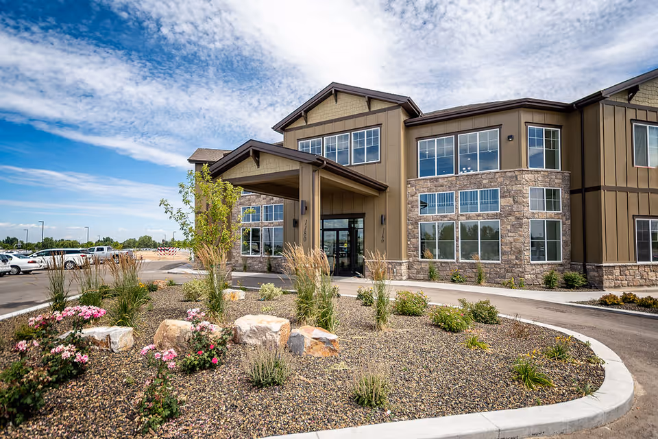 Exterior view of Spring Gardens Senior Living Meridian building with a covered entrance, large windows, stone and brown panel siding, landscaped area with rocks, flowers, and shrubs in front, and a parking lot to the left under a partly cloudy sky.