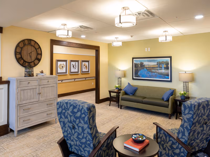 A cozy senior living facility common area with a green sofa adorned with blue pillows, two blue patterned armchairs, a round table with books and a bowl, two side tables with lamps and plants, a large wall clock, and framed pictures of dogs on the wall in the hallway.