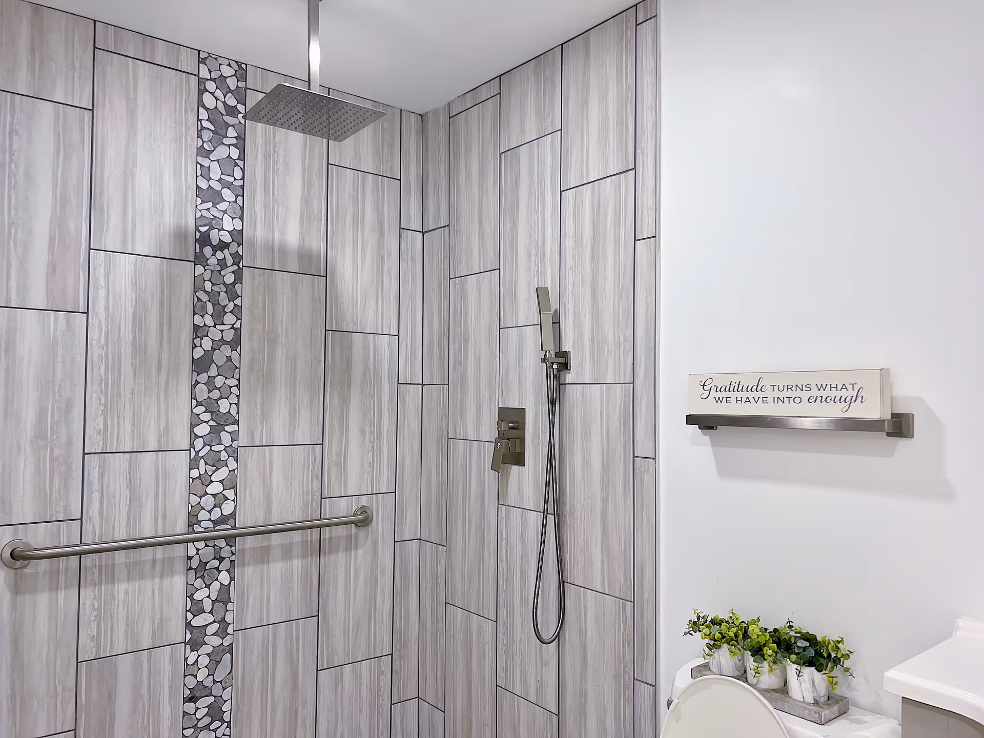 Modern shower area with large rectangular gray tiles and a vertical strip of pebble-like stones. The shower features a square overhead rain showerhead, a handheld showerhead, and a horizontal grab bar. To the right, a white wall has a small shelf holding a decorative sign that reads 'Gratitude turns what we have into enough' and a tray with small green plants. Part of a white toilet and a sink countertop are visible.