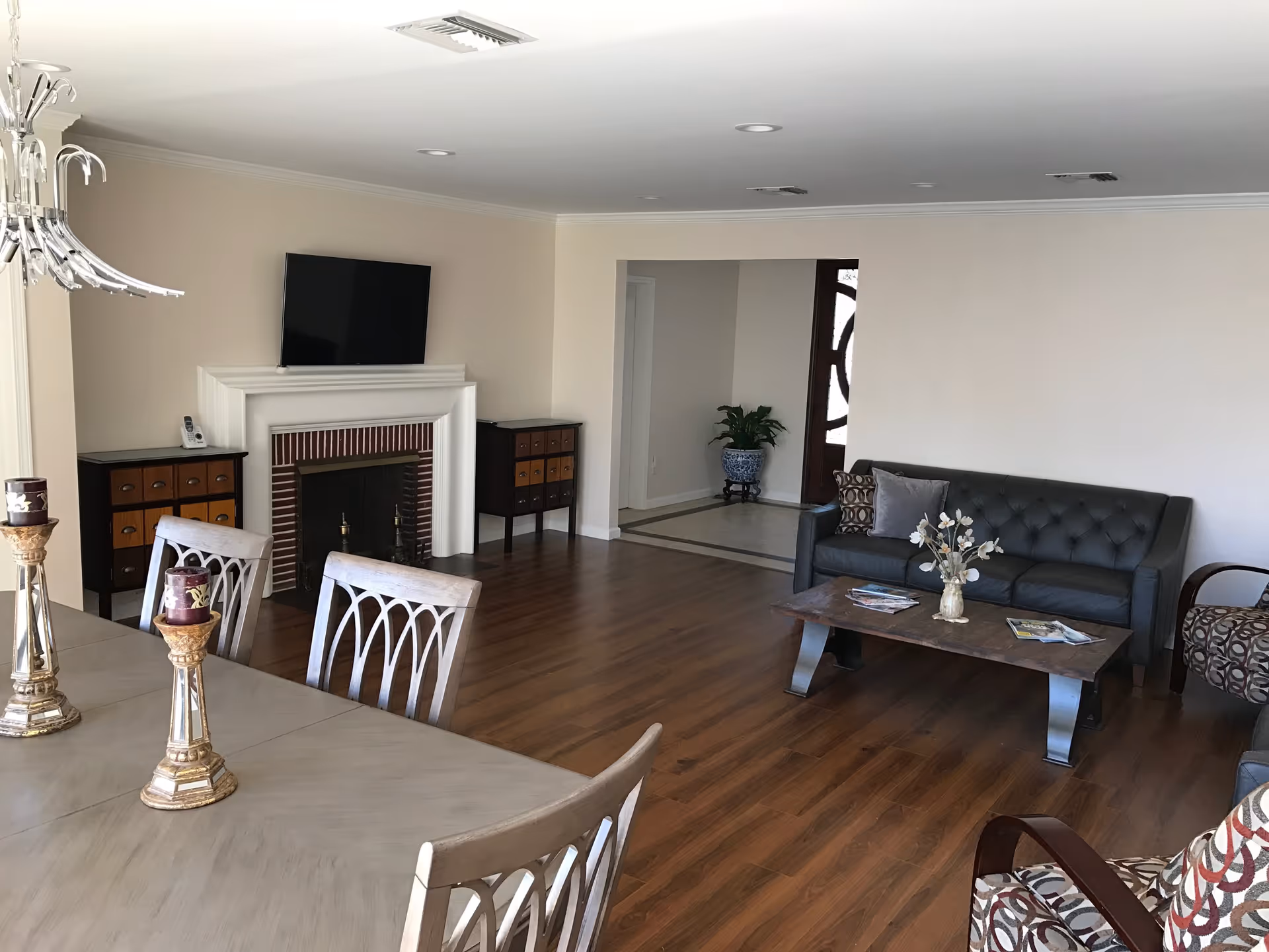 A spacious living and dining area with wooden flooring. The living room features a dark gray tufted sofa, a patterned armchair, a wooden coffee table with a vase of flowers and magazines. The dining area has a light-colored wooden table with matching chairs and decorative candle holders. A white fireplace with a mounted flat-screen TV is visible, flanked by two small cabinets. The walls are painted light beige, and there is a doorway leading to another room with a plant in a blue and white pot.