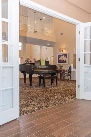 Open double doors lead into a spacious senior living common room featuring a grand piano, seating, patterned carpet, and a second-floor balcony.