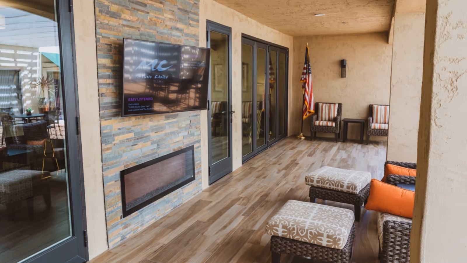 A covered outdoor patio area with wooden flooring, a mounted flat-screen TV above a modern fireplace on a stone accent wall, wicker chairs with orange cushions, patterned ottomans, and two striped cushioned chairs with a small table near an American flag in the corner.