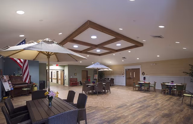 Spacious indoor common area with multiple tables and chairs arranged for seating. Some tables have large umbrellas over them. The room has wooden flooring, a ceiling with recessed lighting and wooden beams, and an American flag displayed near a piano on the left side. There are doors and hallways visible in the background.