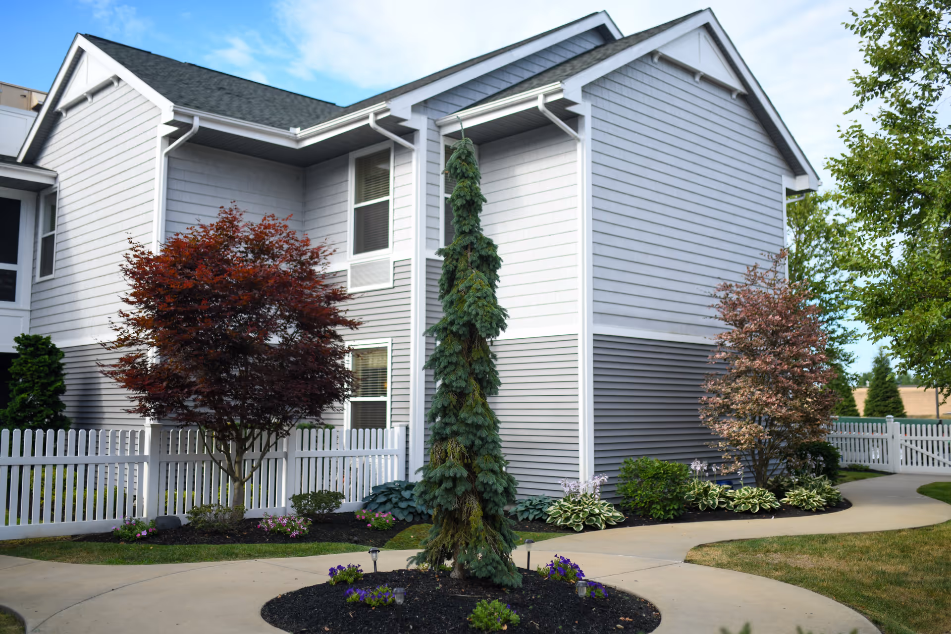 Exterior view of a senior living facility building with gray siding and white trim, surrounded by a white picket fence, landscaped with various trees and shrubs, and a curved concrete walkway.