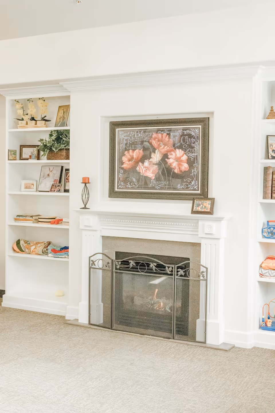 A cozy living room area featuring a white fireplace with a decorative black metal screen. Above the fireplace hangs a framed floral painting with pink flowers and decorative text. On either side of the fireplace are built-in white shelves displaying various framed photos, plants, books, and decorative items. The room has a light-colored carpet and walls, creating a bright and inviting atmosphere.