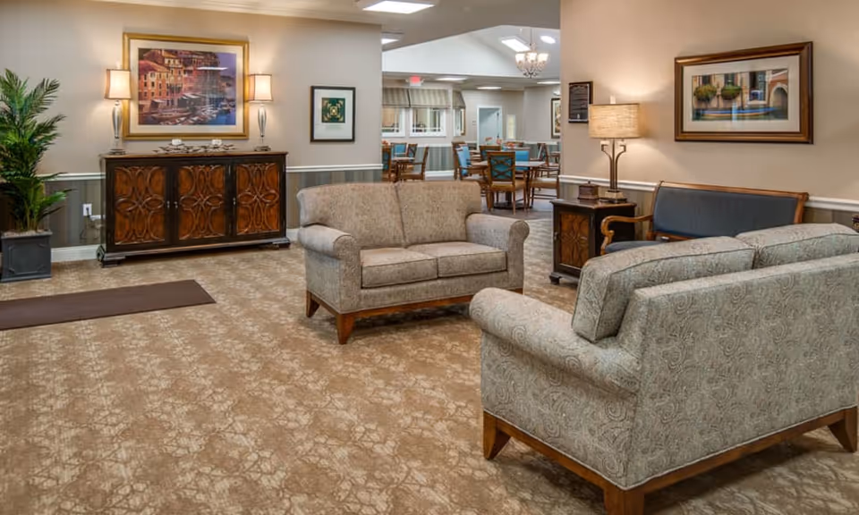 A cozy senior living common area with two patterned sofas and a wooden bench with blue cushions. There is a wooden side table with a lamp and framed artwork on the walls. In the background, a dining area with tables and chairs is visible under a chandelier. A decorative wooden cabinet and a potted plant are also present.