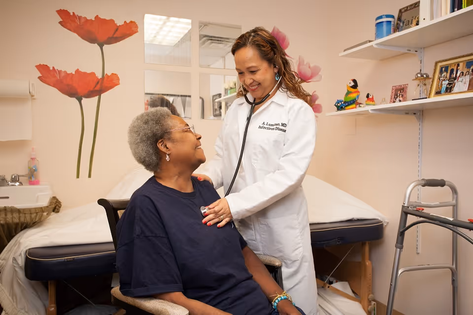 A healthcare professional wearing a white coat uses a stethoscope to listen to the chest of an elderly woman seated in a chair inside a medical examination room. The room has a medical examination table, a walker, shelves with framed photos and decorations, and a wall with large red flower decals.