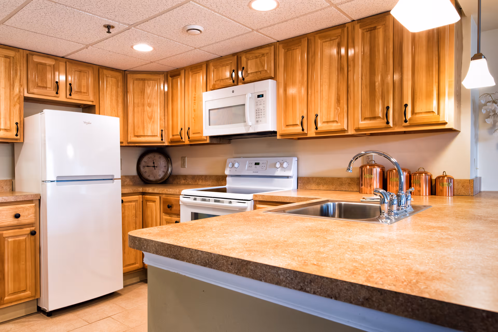 A kitchen with wooden cabinets, a white refrigerator, a white microwave above a white stove, a countertop with a built-in sink and chrome faucet, and copper canisters on the counter. The ceiling has recessed lighting and a hanging pendant light.