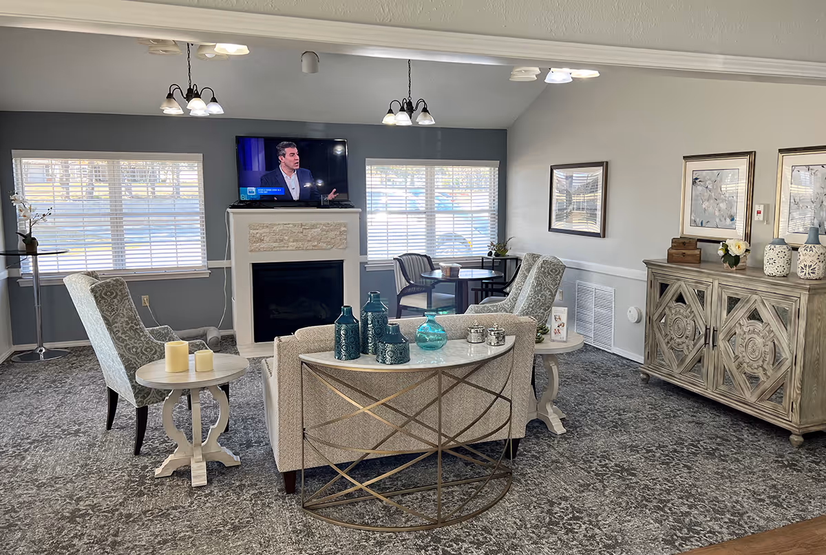 A cozy living room area in Broadmoor Court featuring a fireplace with a TV mounted above it, two patterned armchairs, a beige sofa with decorative vases on a console table behind it, a side table with candles, a wooden cabinet with decorative items, and large windows with blinds letting in natural light.