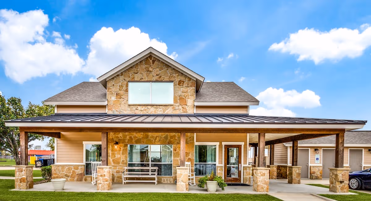 Front exterior view of a two-story senior living facility building with stone and beige siding, a covered porch with wooden pillars, benches, rocking chairs, potted plants, and a clear blue sky with some clouds.