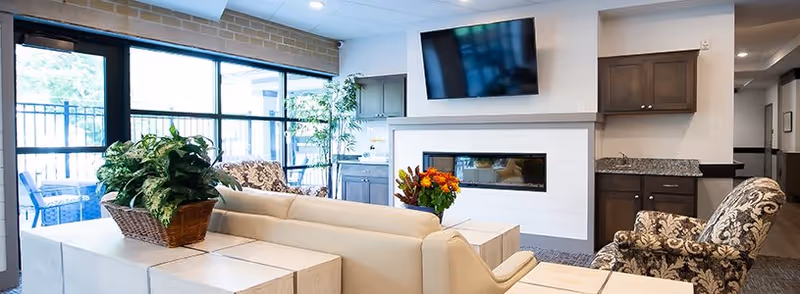 A bright and modern senior living common area featuring a beige sofa, patterned armchairs, a fireplace with a mounted TV above it, large windows allowing natural light, indoor plants, and a small kitchenette with dark cabinets and granite countertops.
