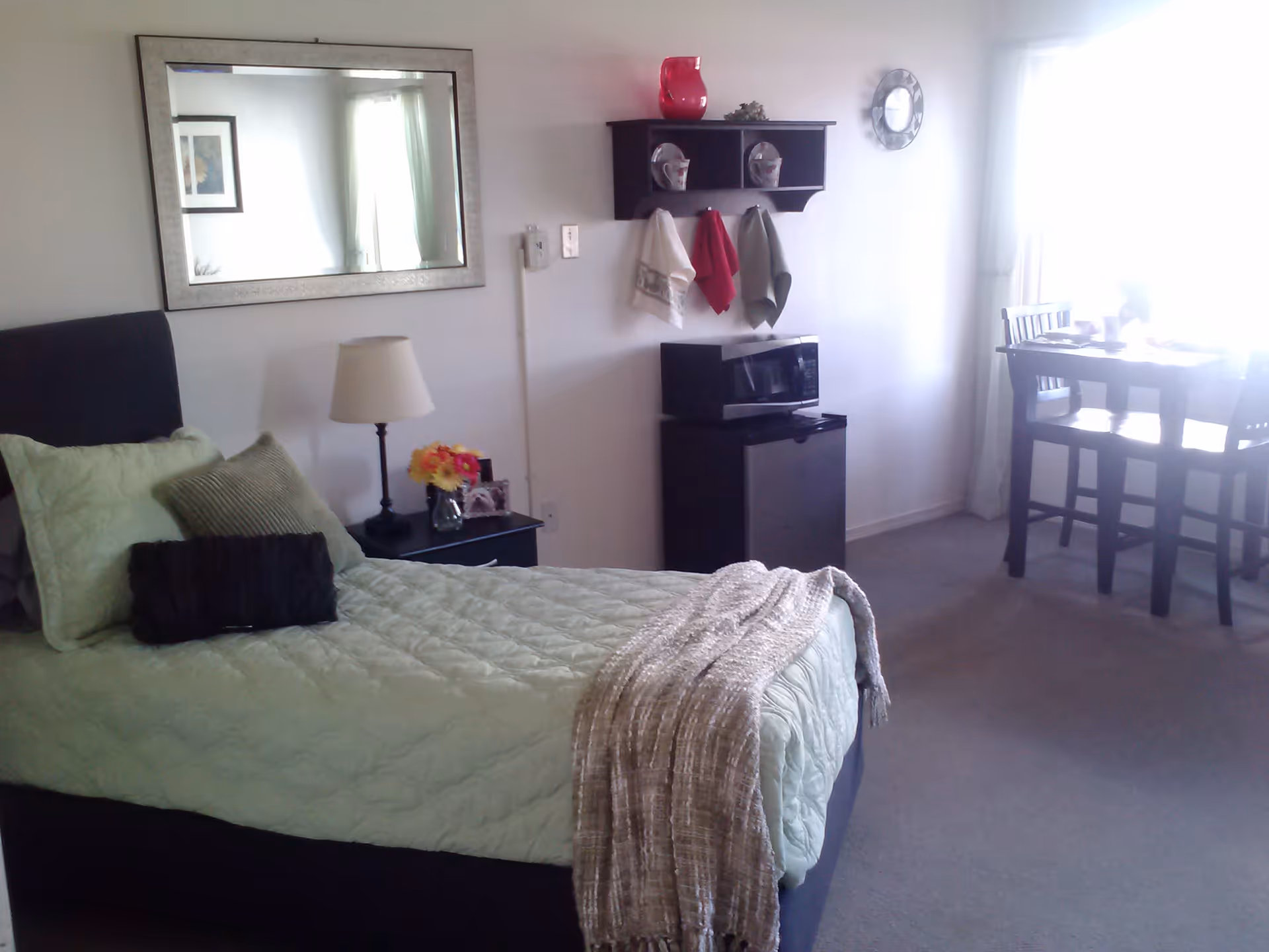 A cozy bedroom with a single bed made with light green bedding and multiple pillows. Next to the bed is a small black nightstand with a lamp, a small vase of flowers, and a framed photo. Above the nightstand is a large rectangular mirror. On the opposite wall, there is a small black shelf holding decorative items and three hanging towels. Below the shelf is a black microwave on top of a small refrigerator. In the background, there is a dining table with two chairs near a window with white curtains letting in bright light.