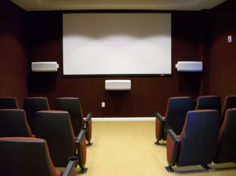 Small theater room with rows of black and red cushioned seats facing a large white projection screen mounted on a dark brown wall, with three white speakers attached to the wall around the screen.