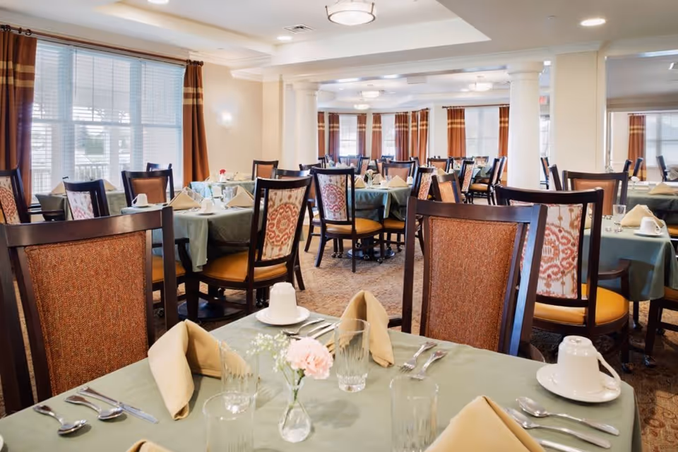 A spacious dining room with multiple tables covered in green tablecloths, each set with beige napkins, glassware, silverware, and white cups. The room features wooden chairs with patterned upholstery, large windows with brown curtains, and soft ceiling lighting.