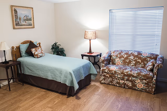 A cozy bedroom with a single bed covered in a light blue quilt and decorative pillows. Next to the bed is a small wooden side table with a lamp and a plant. Across from the bed is a floral-patterned loveseat under a window with closed blinds. A framed painting hangs on the wall above the bed.