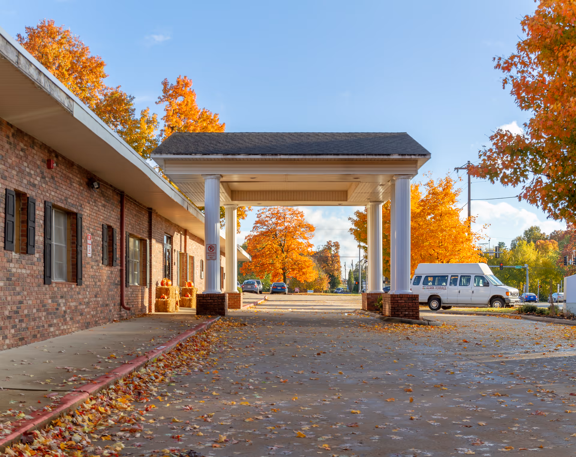 Exterior view of The Blossoms at Rogers Rehab & Nursing Center showing a brick building with a covered entrance supported by white columns. The driveway and surrounding area are covered with fallen autumn leaves, and colorful fall trees are visible in the background along with a white van parked nearby.