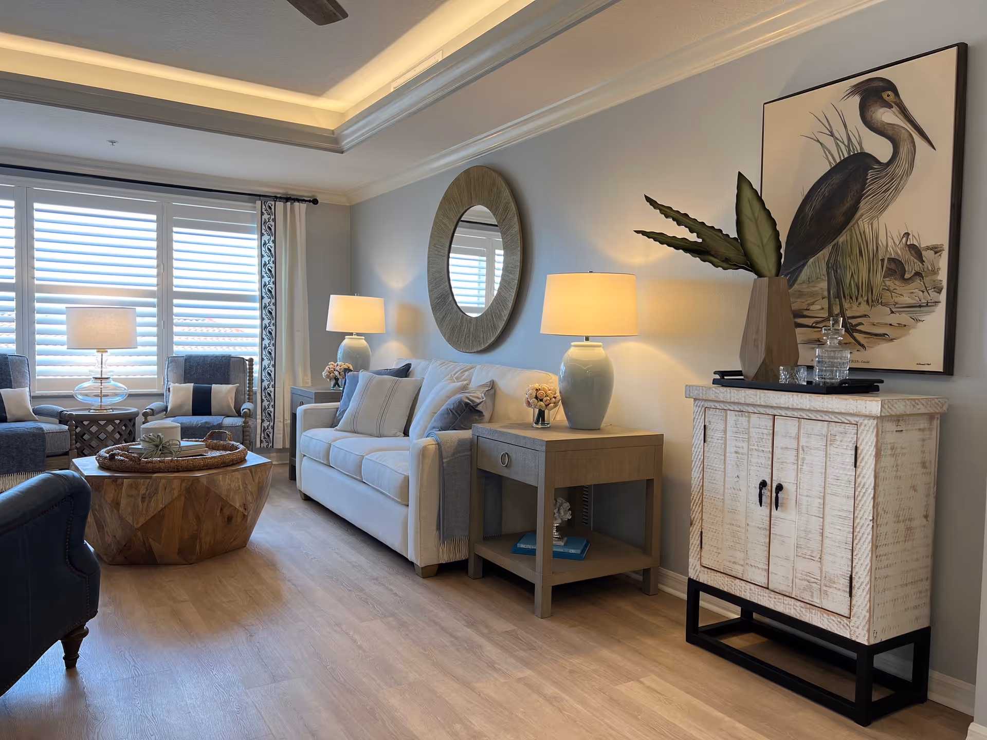 Light-filled living room with a sofa, two armchairs, wooden coffee table, side tables with lamps, a round mirror and bird artwork on the wall.
