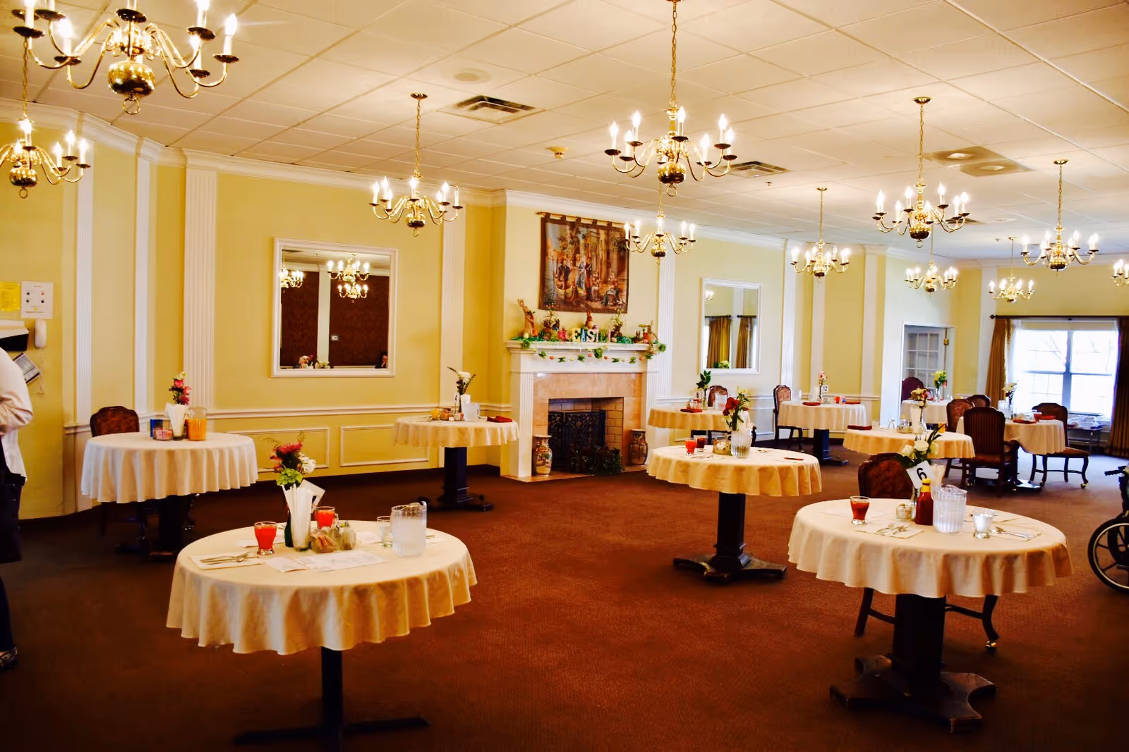 A spacious dining room with round tables covered in white tablecloths, each set with glasses, pitchers, and small flower arrangements. The room features yellow walls, multiple chandeliers hanging from the ceiling, a fireplace with decorative items above it, and large windows letting in natural light.