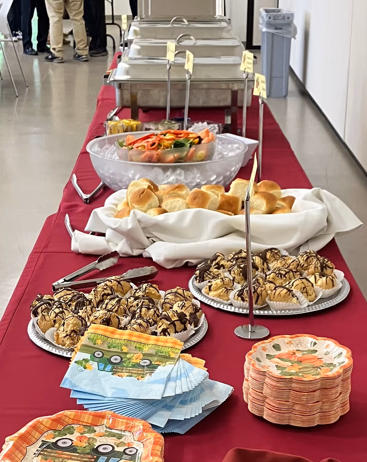 A buffet table covered with a red tablecloth displaying trays of food including bread rolls, a bowl of salad on ice, and dessert pastries drizzled with chocolate. There are also stacks of decorative napkins and small plates with a fall-themed design. In the background, people are partially visible standing near the end of the table.