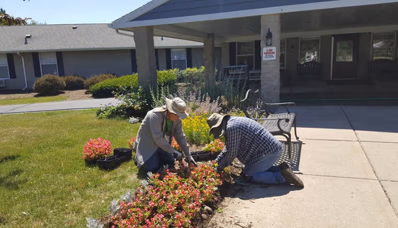 Two people wearing hats are kneeling and planting flowers in a garden bed outside a building with a covered entrance. There are benches nearby and a sign on a pillar that says 'No Smoking Within 50 Feet'.