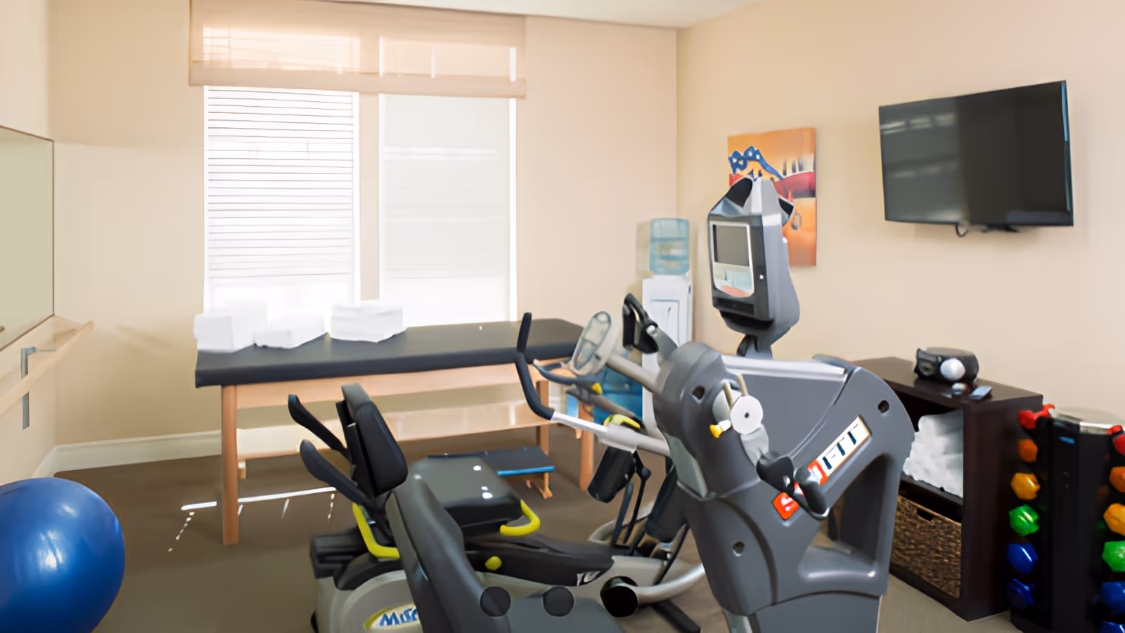 A small fitness room with exercise equipment including a recumbent bike and elliptical machine, a blue exercise ball, a massage or therapy table with folded towels, a water cooler, a wall-mounted TV, a rack of colorful dumbbells, and a small cabinet with towels and a radio.