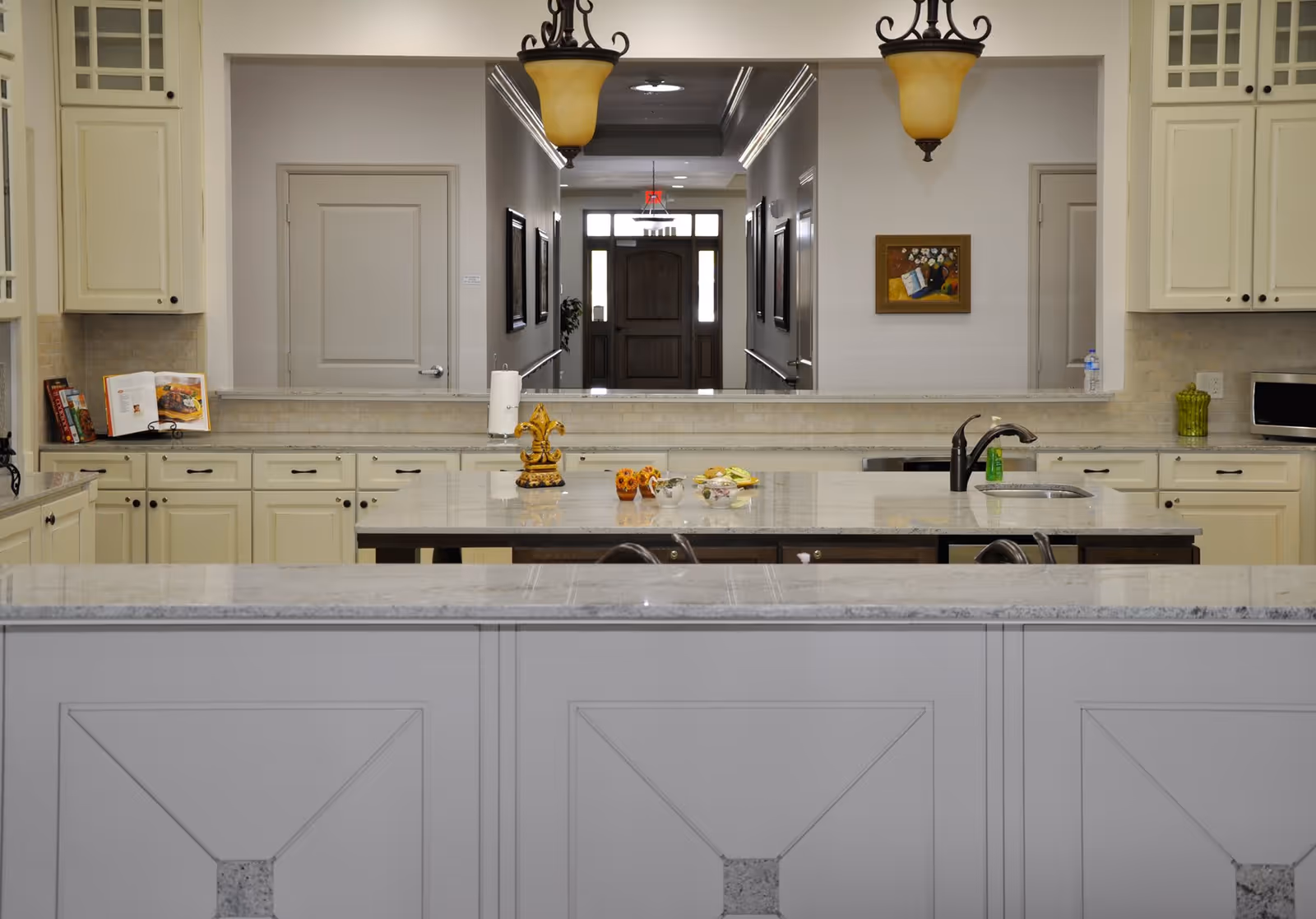 A spacious kitchen with cream-colored cabinets, a large island countertop with a sink and faucet, decorative items on the island, and two hanging pendant lights. In the background, there is a hallway leading to a wooden front door with glass panels on either side. The kitchen has a clean and elegant design with granite countertops and a microwave on the right side.