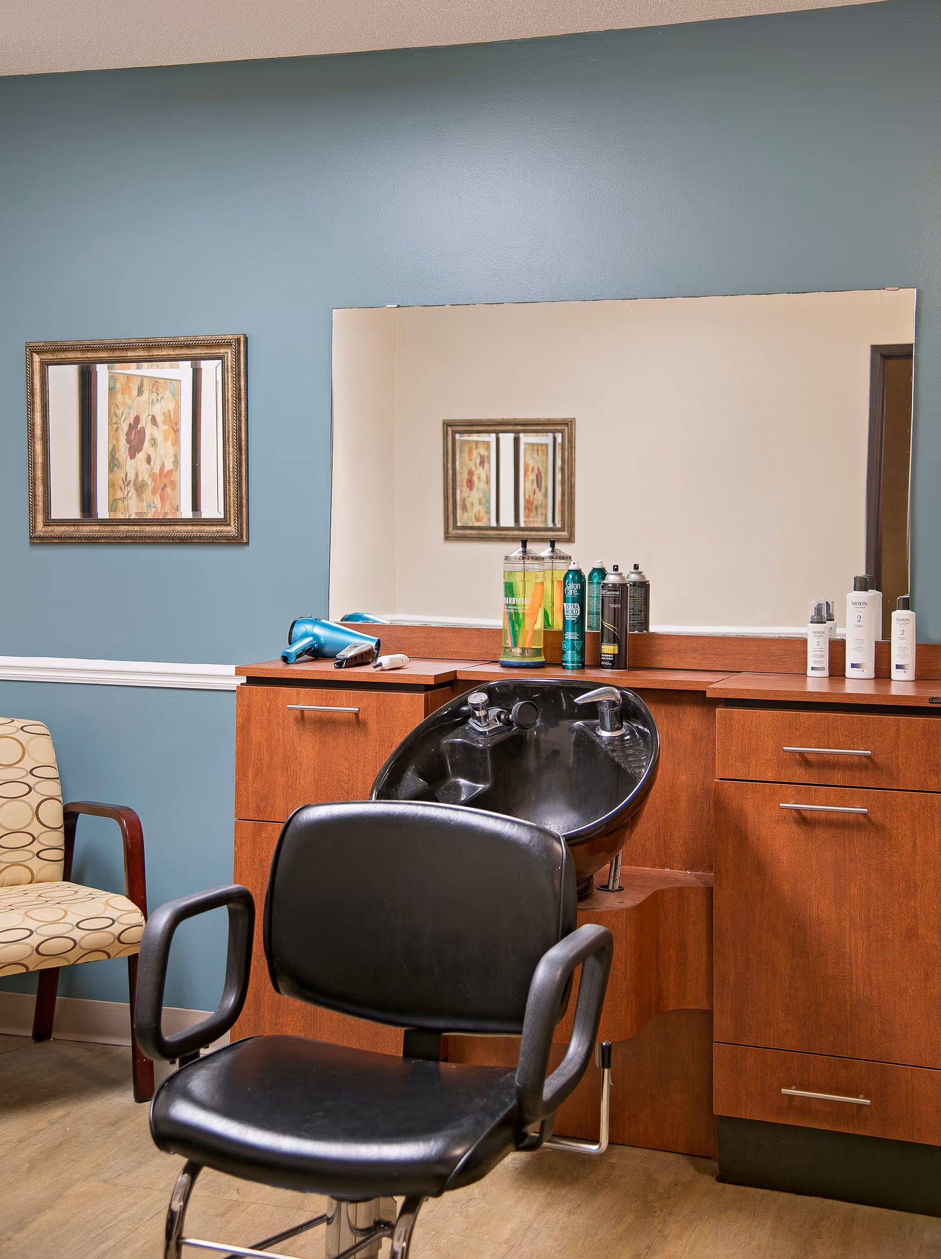 Interior view of a hair salon area in a senior living facility with a black salon chair in front of a black hair washing sink. The wooden counter behind the sink holds various hair care products and a blue hair dryer. A large mirror is mounted on the wall above the counter, reflecting a framed floral artwork on the opposite wall. There is also a beige chair with a circular pattern on the left side of the image.