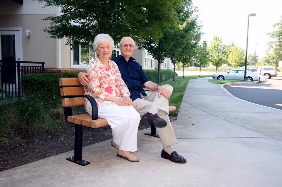 An elderly couple sitting on a wooden bench outside near a retirement community building. The woman is wearing a floral blouse and white pants, and the man is wearing a dark blue shirt and beige pants. They are smiling and sitting close together with trees and parked cars visible in the background.
