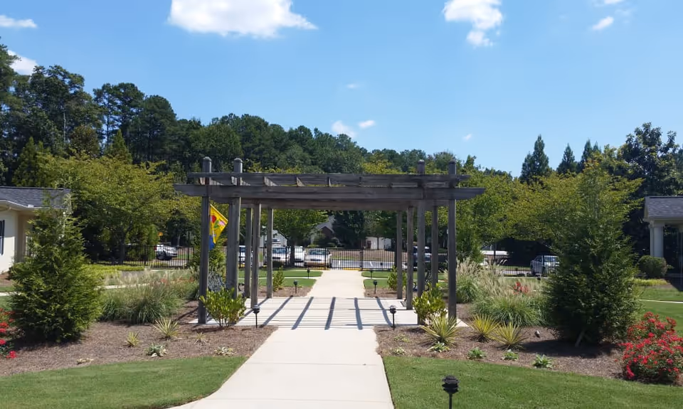 A landscaped concrete walkway with a wooden pergola leading to a gated entrance surrounded by trees, shrubs, and buildings.