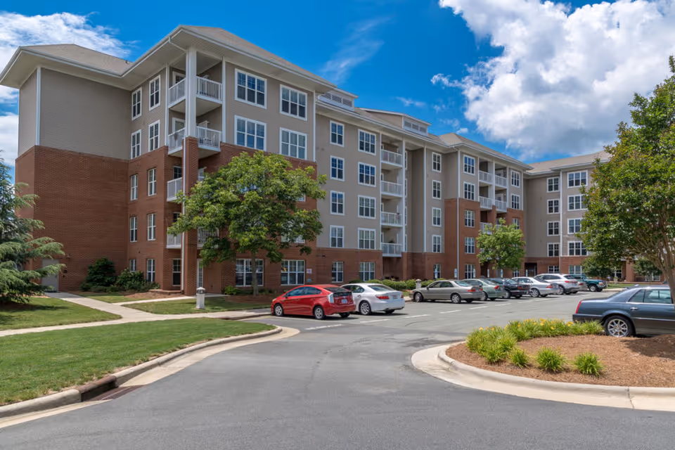 Large multi-story senior living building with balconies, a parking lot with cars, landscaped islands, and a partly cloudy blue sky.