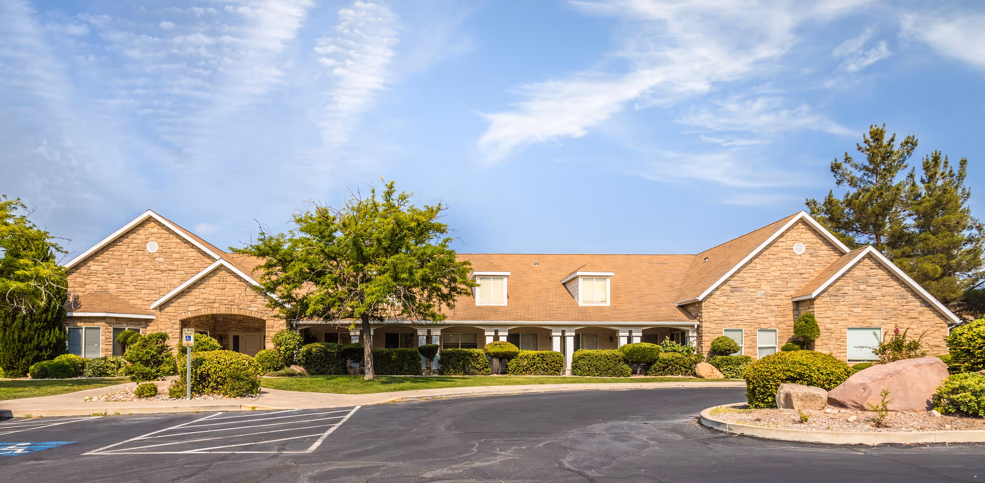 Exterior front view of a single-story assisted living facility building with a tan brick facade, multiple windows, a covered entrance, and a landscaped area with trees and bushes under a partly cloudy sky.