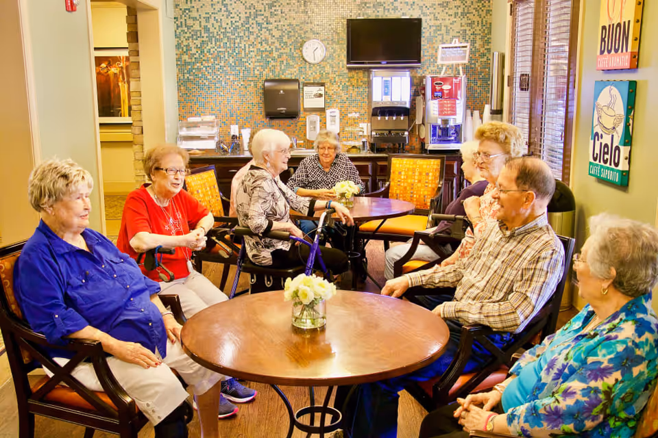 A group of elderly people sitting around two wooden tables in a cozy common area with mosaic tile wall, a clock, a TV, and beverage machines in the background. The room has warm lighting and decorative signs on the walls.